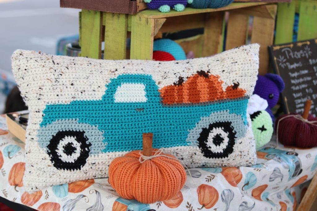 White crochet pillow of blue truck with three orange pumpkins in tailgate